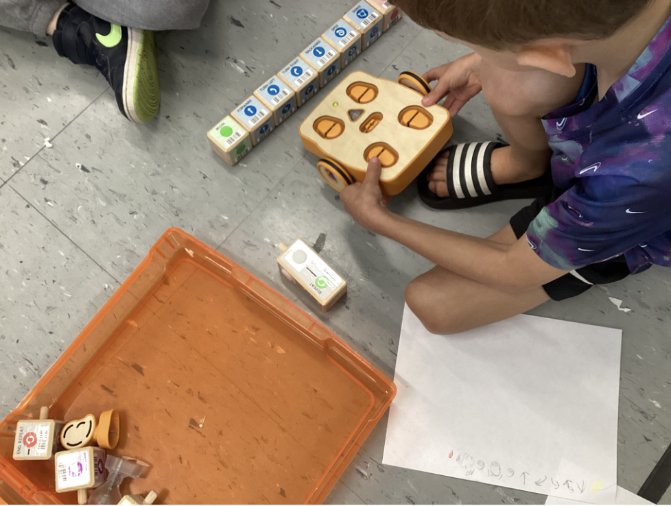 A student sits on the floor, working with a KIBO robot and coding blocks. 